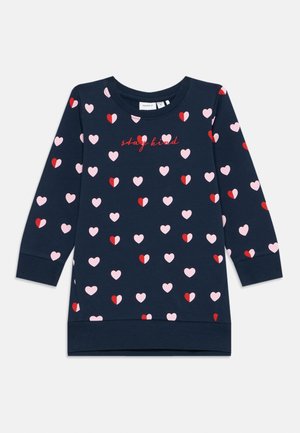 Navyblå kjole med lange ærmer med lyserøde og røde hjerte mønstre og rød tekst "stay kind" på brystet.