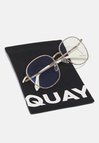 Occhiali da sole oversize in tonalità oro con lenti grigio scuro sfumate, dotati di un bordo testurizzato, posati su una custodia nera con la scritta "QUAY" in bianco.