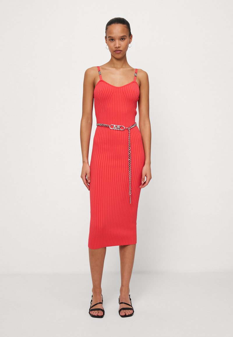 Eine Frau steht in einem eng anliegenden, roten Ripp-Midikleid mit Spaghetti-Trägern und einem silbernen Kettengürtel, kombiniert mit schwarzen Open-Toe-Absätzen.