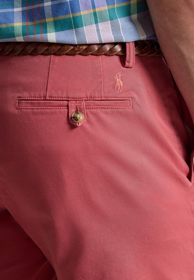 Ανδρικά ροζ chino σορτς με πίσω τσέπη, κούμπωμα με κουμπί και διακριτική κεντημένη λογότυπο. Υφή υφάσματος και πλεξούδα καφέ ζώνη εμφανής.