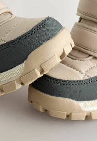 Beige og grå vandresko med en tekstureret overflade, polstret overdel, velcro-remme og robuste såler for godt greb.