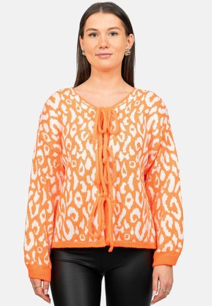 Woman wearing an orange and white leopard print knit sweater with front ties, paired with black pants, standing against a plain background.