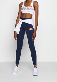 Crop top sportivo bianco con scritte grigie, abbinato a leggings navy con un logo multicolore. Sneakers bianche completano l'outfit.