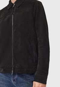 Person iklædt en sort suede zip-up jakke med krave og sidelommer, parret med blå denim jeans på en ensfarvet baggrund.
