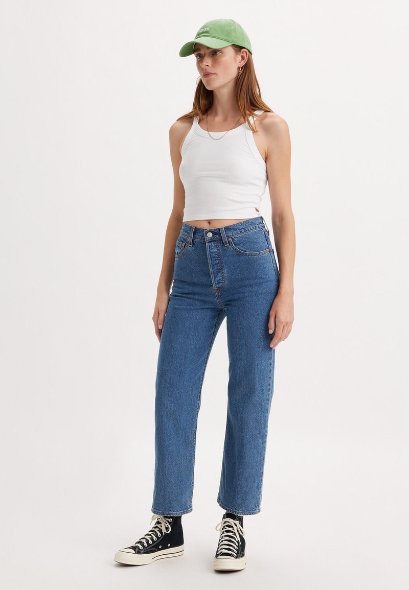 Top court blanc, jeans taille haute bleus, baskets noires et blanches, et casquette vert clair. La tenue présente une coupe décontractée et confortable.