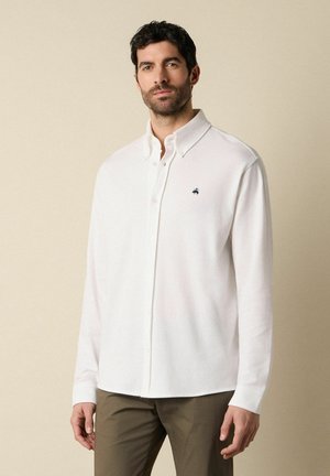 Muž s tmavými kudrnatými vlasy, oblečený v bílé košili na knoflíky s malým černým logem a olivově zelených kalhotách, stojící na béžovém pozadí.