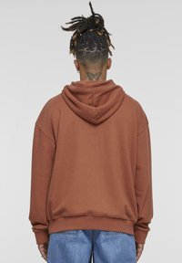 Sudadera marrón de gran tamaño con capucha ajustable, puños y dobladillo acanalados, y una textura suave. Llevada sobre jeans de color azul claro.
