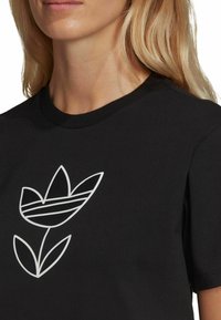 T-shirt in cotone nero con una grafica di un fiore e foglie in outline bianco, dotata di dettagli a righe orizzontali sui petali del fiore.