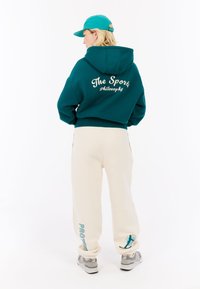 Tyrkysový skrátený mikina s bielym nápisom na chrbte, spárovaná s krémovými nohavicami so tyrkysovým textom a akcentmi, a sivými teniskami.