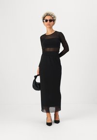 Schwarzes langärmliges Kleid mit durchsichtigen Einsätzen, einer taillierten Taille und einem locker fallenden Rock. Kombiniert mit schwarzen spitzen High Heels und einer kleinen schwarzen Handtasche.