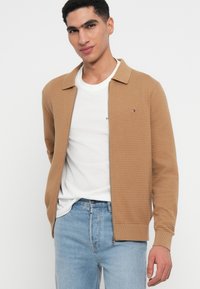 Barna kötött, zipzáros dzseki, galléros nyakkal, bordázott mandzsettákkal és finom vízszintes textúrával. Fehér póló és világos farmernadrág fölött viselve.