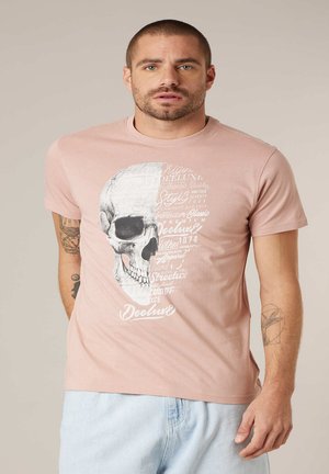 Homme portant un t-shirt rose clair avec un dessin de crâne noir et blanc et du texte blanc, assorti à un jean bleu clair, se tenant devant un fond uni.