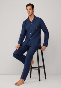 Conjunto de pijama azul marino de tejido suave, que incluye una camisa con botón en la parte delantera y un bolsillo en el pecho, además de un pequeño diseño bordado, acompañado de pantalones a juego.