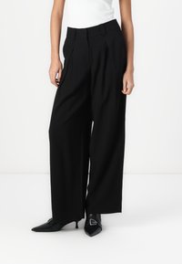 Black wide-leg trousers made of soft fabric, featuring front pleats and belt loops, paired with pointed black shoes.