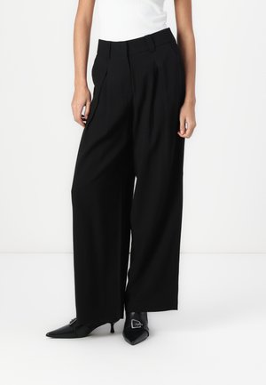 Schwarze weit geschnittene Hosen aus weichem Stoff, mit vorderen Falten und Gürtelschlaufen, kombiniert mit spitzen schwarzen Schuhen.