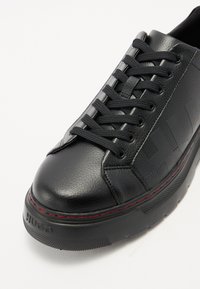 Baskets en cuir noir avec une tige texturée, bout rond, lacets plats, coutures rouges, et une semelle plateforme, arborant un logo en relief sur le côté.