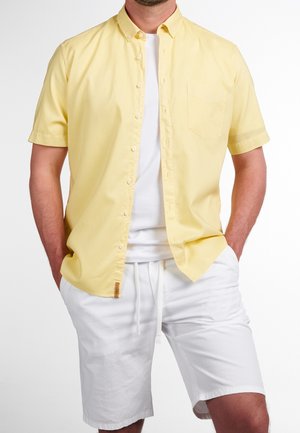 Mann trägt ein aufgeknöpftes gelbes Kurzarmhemd über weißem T-Shirt mit weißen Shorts mit Kordelzug, Hände in den Taschen, vor einfarbigem Hintergrund.