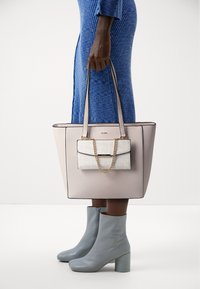 Velká béžová taška s tmavým lemováním, ve které je menší bílá taška s řetízkem. Modelka má na sobě světle modré kotníkové boty a strukturované modré šaty.