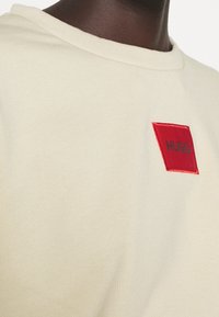 Kremowa bluza z ribbowym dekoltem i czerwoną prostokątną naszywką z napisem "HUGO" czarnym haftem. Miękka tekstura materiału.