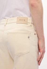 Jean en denim beige avec une étiquette en cuir "BOSS" au-dessus de la poche arrière et un petit logo métallique "B" près du bord de la poche.