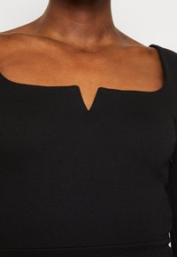 Musta pitkähihaista toppi, jossa on neliönmuotoinen pääntie ja keskellä V-muotoinen aukko. Valmistettu sileästä ja joustavasta kankaasta.