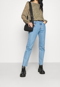 Langärmlige olivgrüne Bluse mit Blumenmuster, hellblaue gerade geschnittene Jeans mit ausgefranstem Saum, schwarze Stiefeletten, kleine schwarze Umhängetasche.