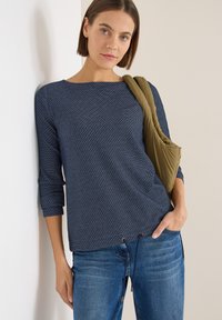 Navy top w wzór marynarski z rękawami o długości trzech czwartych, okrągłym dekoltem i regulowanym sznurkiem u dołu, w połączeniu z niebieskimi dżinsami i oliwkową torbą.