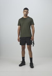 Camiseta verde oliva y pantalones cortos negros, ambos de tejido transpirable. Zapatos y calcetines negros con un logo. Sosteniendo una cuerda para saltar, fondo neutro.