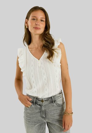 Mujer con cabello largo y castaño vistiendo una blusa blanca sin mangas con volantes y vaqueros grises de cintura alta sobre un fondo gris liso.