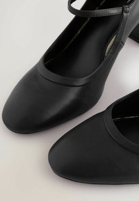 Zwarte leren pumps met een ronde neus en een fijne enkelband. Soepele textuur met subtiele stikseldetails langs de randen.