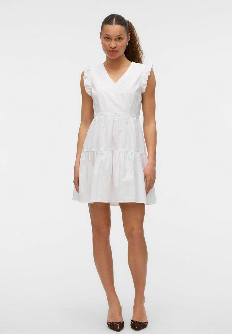 Femme aux cheveux bouclés portant une robe blanche sans manches à col en V à étages et des ballerines foncées à bout pointu, debout devant un fond uni.
