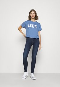 Mėlynas medvilninis marškinėlis su baltu "LEVI'S" logotipu, derinamas su tamsiai mėlynais lieknėjimo džinsais ir baltais drobės sportbačiais. Modelis stovi tiesiai.