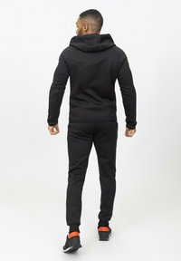 Sweatshirt noir avec capuche et jogging assorti, présentant un design ajusté et des poignets côtelés. Accentué de détails orange sur les chaussures.