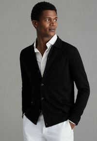 Cardigan negru cu nasturi, purtat peste o cămașă albă cu guler. Are mâneci lungi și un design simplu, asortat cu pantaloni albi.