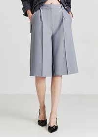 Personne portant des shorts gris à jambes larges de longueur genou avec des plis, une veste noire rayée et des chaussures à talons aiguilles noires, les mains dans les poches.