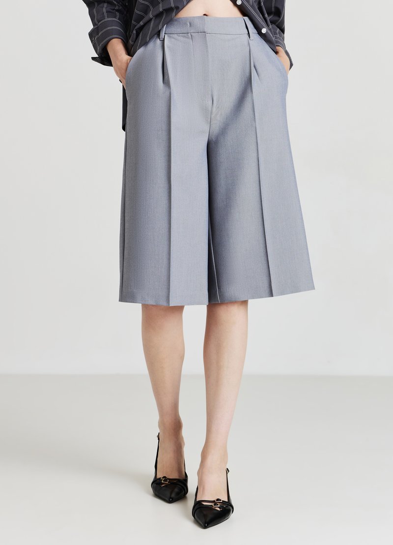 Personne portant des shorts gris à jambes larges de longueur genou avec des plis, une veste noire rayée et des chaussures à talons aiguilles noires, les mains dans les poches.