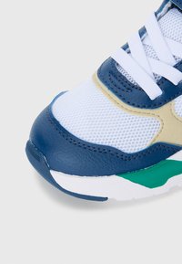 Zapatilla deportiva con parte superior de cuero y malla azul, acentos beige, cordones blancos y detalles en la suela verde. Acabado texturizado visible.