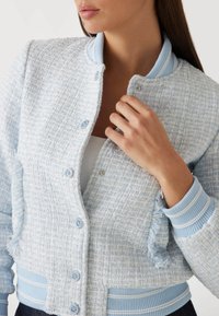 Bomber jacket texurizzato di colore azzurro chiaro con polsini e colletto a coste. Presenta chiusure a bottone e dettagli in frange sulle maniche.