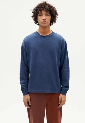 Junge Person mit lockigem Haar, die ein blaues Langarmhemd und braune Hosen trägt und vor einem schlichten hellen Hintergrund steht.