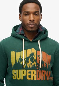 Zielona bluza z kapturem z kolorową grafiką górską oraz napisem "SUPERDRY" w żółtym i pomarańczowym. Widać sznurki i kraciasty kołnierz.