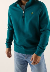 Sini-vihreä vetoketjullinen collegepaita pehmeästä kankaasta, jossa on korkea kaulus ja vasemmassa rinnassa kirjailtu logo; yhdistetty vaaleansinisiiin farkkuihin.