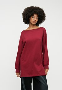 Μπλουζάκι σε μπορντό από oversized μακριά μανίκια με φαρδύ λαιμό σινιέ και ριμπ περιθώρια. Μαλακό βαμβακερό ύφασμα με λεία υφή και πλευρικές σχισμές.