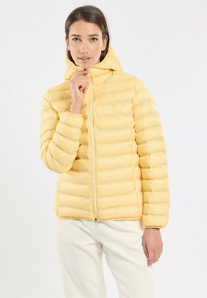 Femme portant une veste matelassée jaune clair avec capuche, légèrement zippée, associée à un pantalon blanc sur un fond blanc uni.