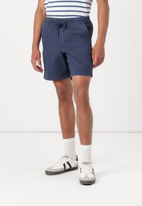 Mørkeblå shorts med elastisk talje og snøre. Har to sidelommer og en lige kant. Bæres med hvide sokker og sneakers.