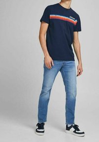 Marinblå T-shirt med ett horisontellt flerfärgat randmönster över bröstet, i kombination med ljusblå jeanstyger och svart-vita sneakers.