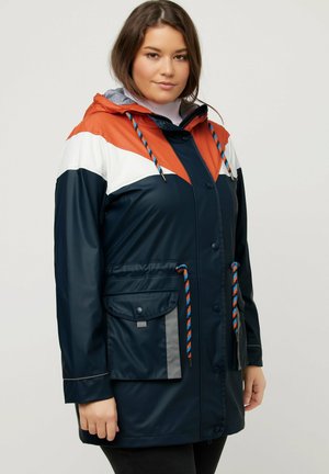 Waterdichte regenjas in marineblauw, oranje en wit. Kenmerken zijn een capuchon, trekkoorddetails en vo pockets met drukknoopsluitingen.