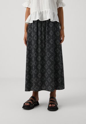 Vrouw met een lange zwart-wit patroonrok, witte blouse met ruches en zwarte plateauzandalen staand op een effen achtergrond.