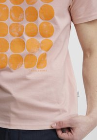 Lys rosa bomull t-skjorte med et design av store, teksturerte oransje sirkler. Teksten sier "CASUAL FRIDAY" under designet.
