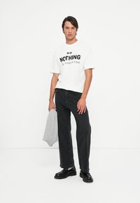 Hvid t-shirt med sort tekst, sorte jeans og sorte sko. En lysegrå sweater draperet over armen. Enkel, afslappet design.