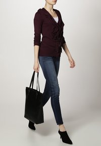 Bordowy sweter rozpinany z falbanami i guzikami, zestawiony z niebieskimi dżinsami i czarną torebką. Modelka nosi czarne szpilki w szpic.